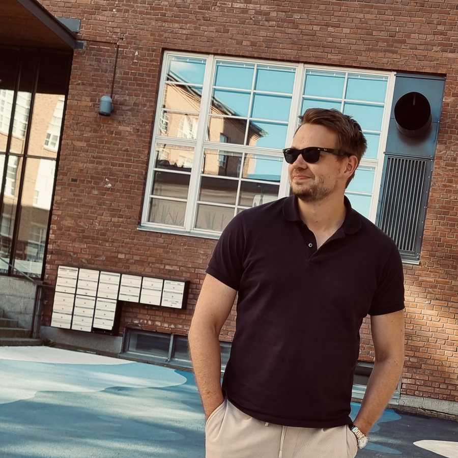 Portrait of Tommi Tuovinen wearing sunglasses and a dark polo shirt, standing in front of a brick building